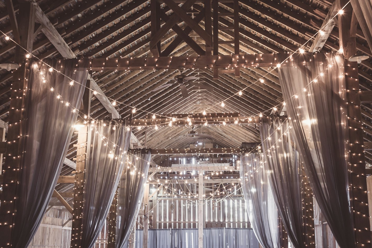 Espace événementiel rustique dans une grange décoré de guirlandes lumineuses et de draperies en tissu.
