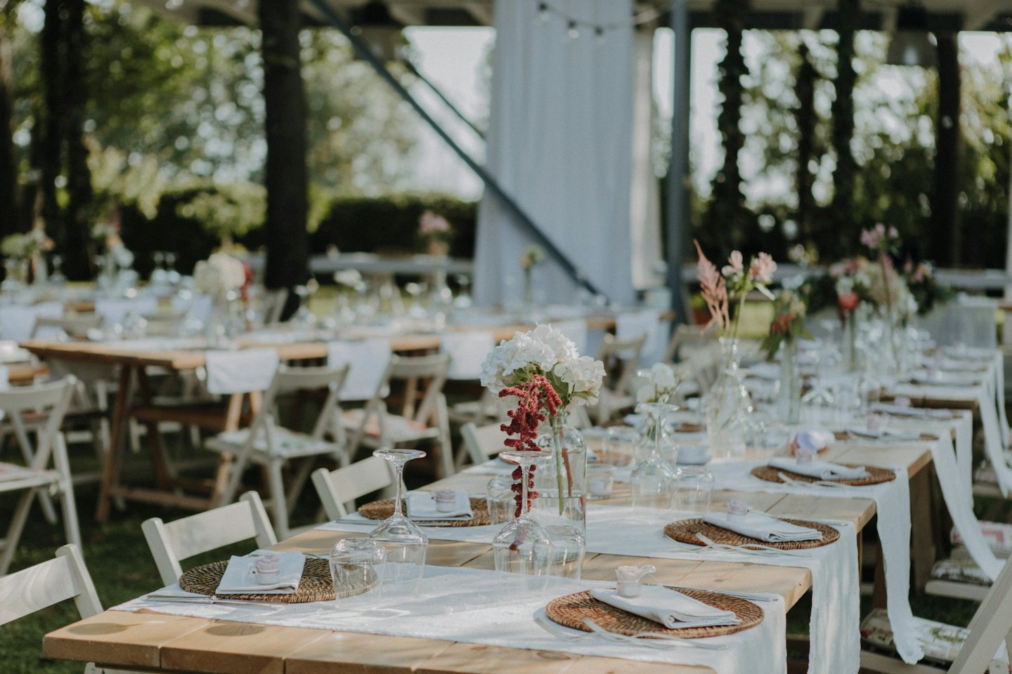Table en bois dressée pour un événement en extérieur, avec des centres de table floraux et des chaises blanches.