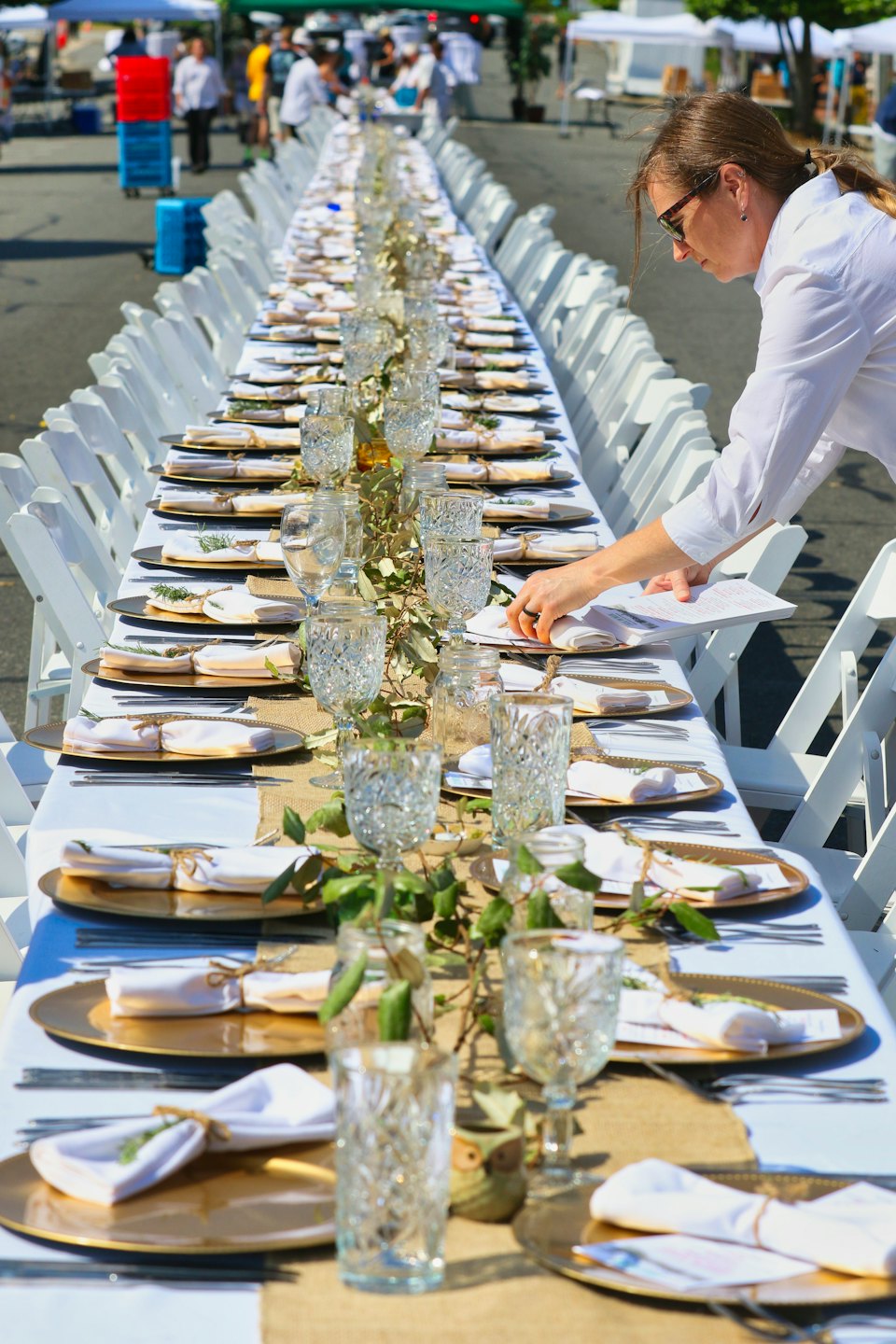 Une organisatrice d'événements dispose les couverts sur une longue table de réception extérieure.